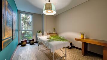 A room with a massage table, a wooden bench, a potted plant, and a large window with a view of greenery.