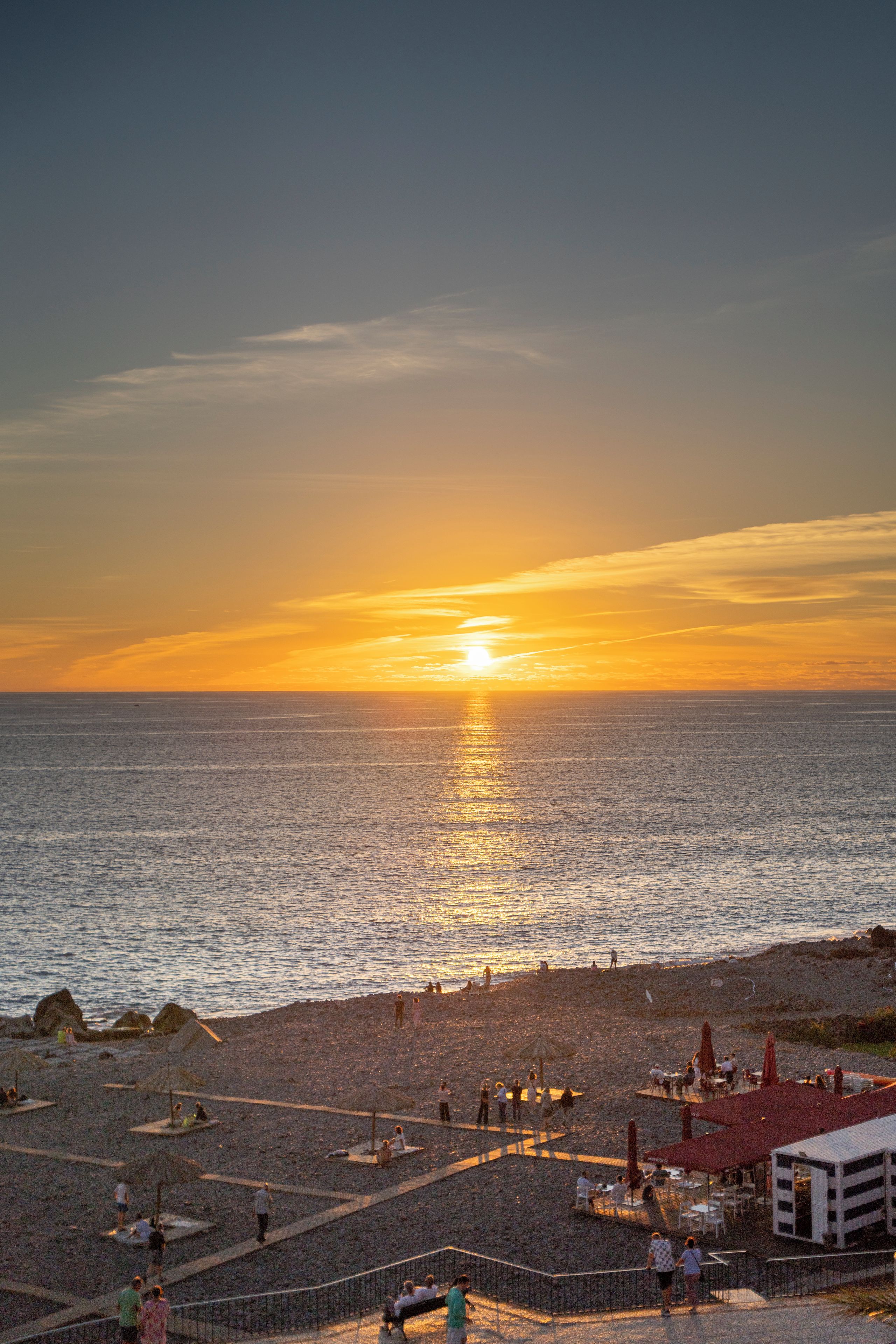 Escena de playa con atardecer, gente y una zona de restaurantes.