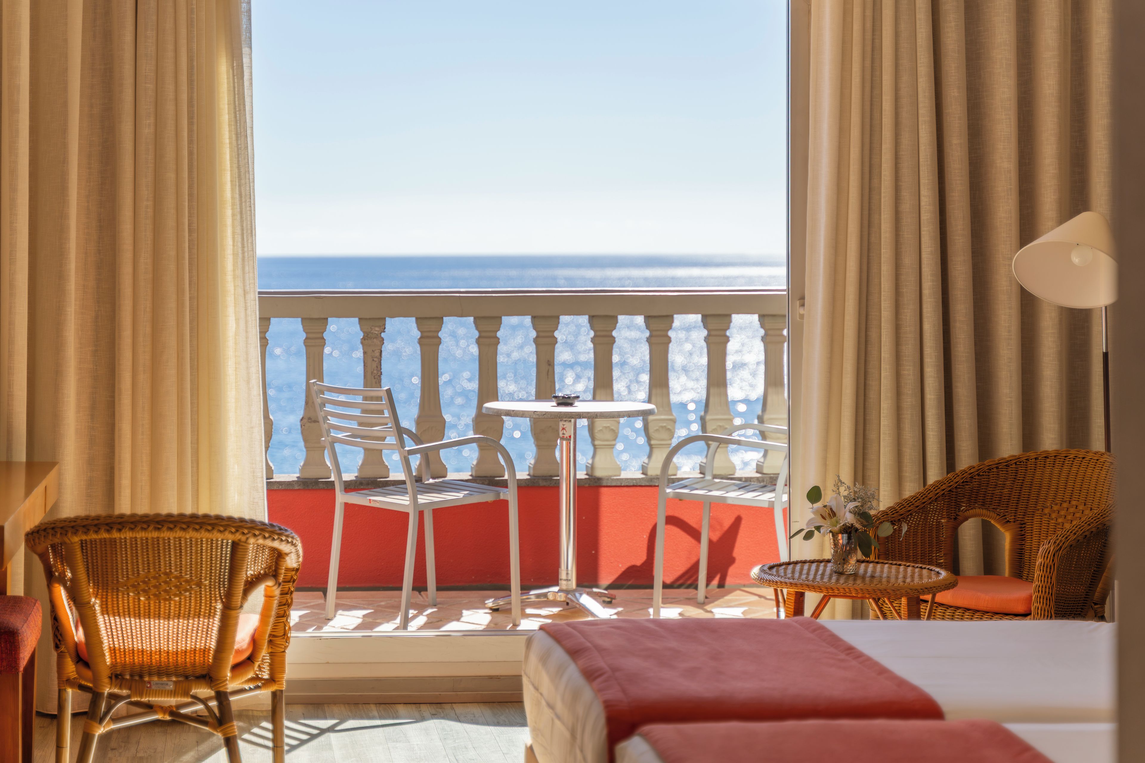 A hotel room with a balcony, two wicker chairs, a small round table, a bed with a red blanket, and a lamp.