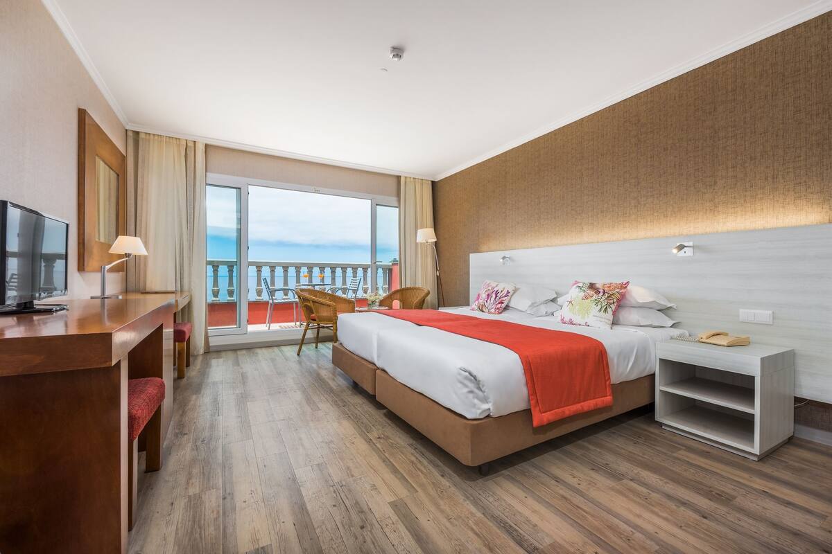 Una habitación de hotel con una cama, un escritorio, una silla, una mesita de noche y un balcón grande con ventanal.