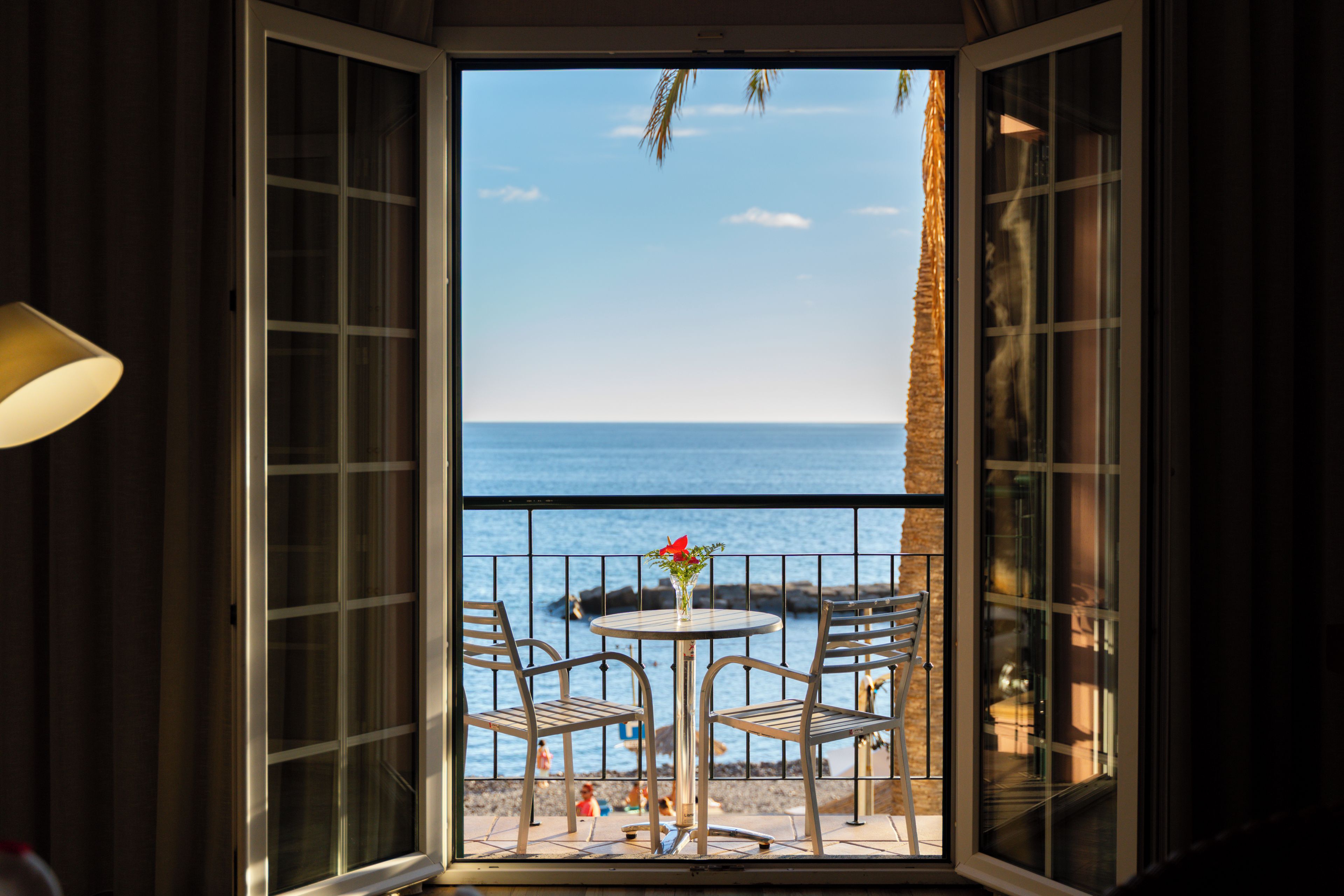 A view from a room with a balcony overlooking a beach, featuring a table with chairs and a vase of flowers.