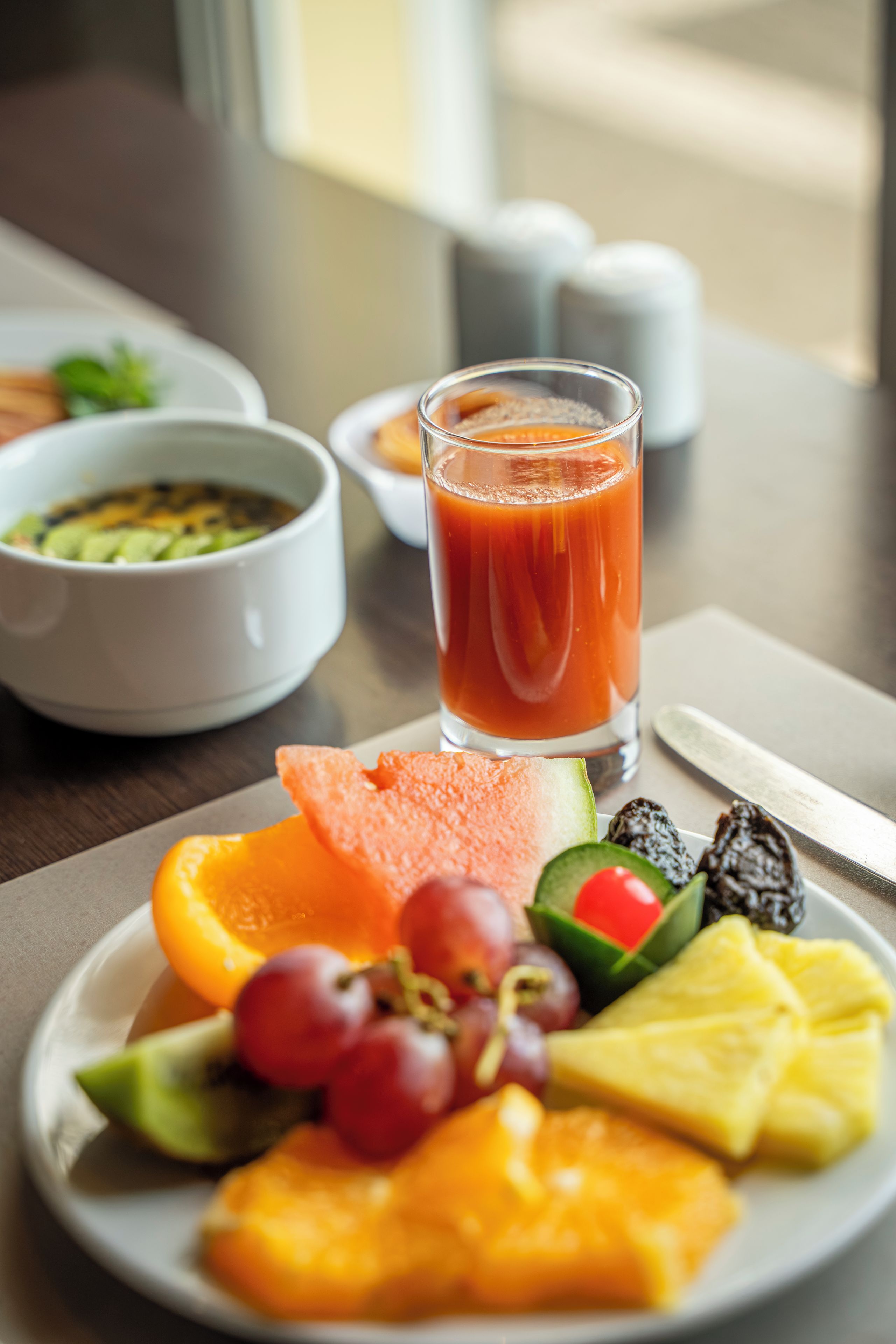 A plate of fruit and a glass of juice on a table with a bowl of soup and a salt shaker in the background.