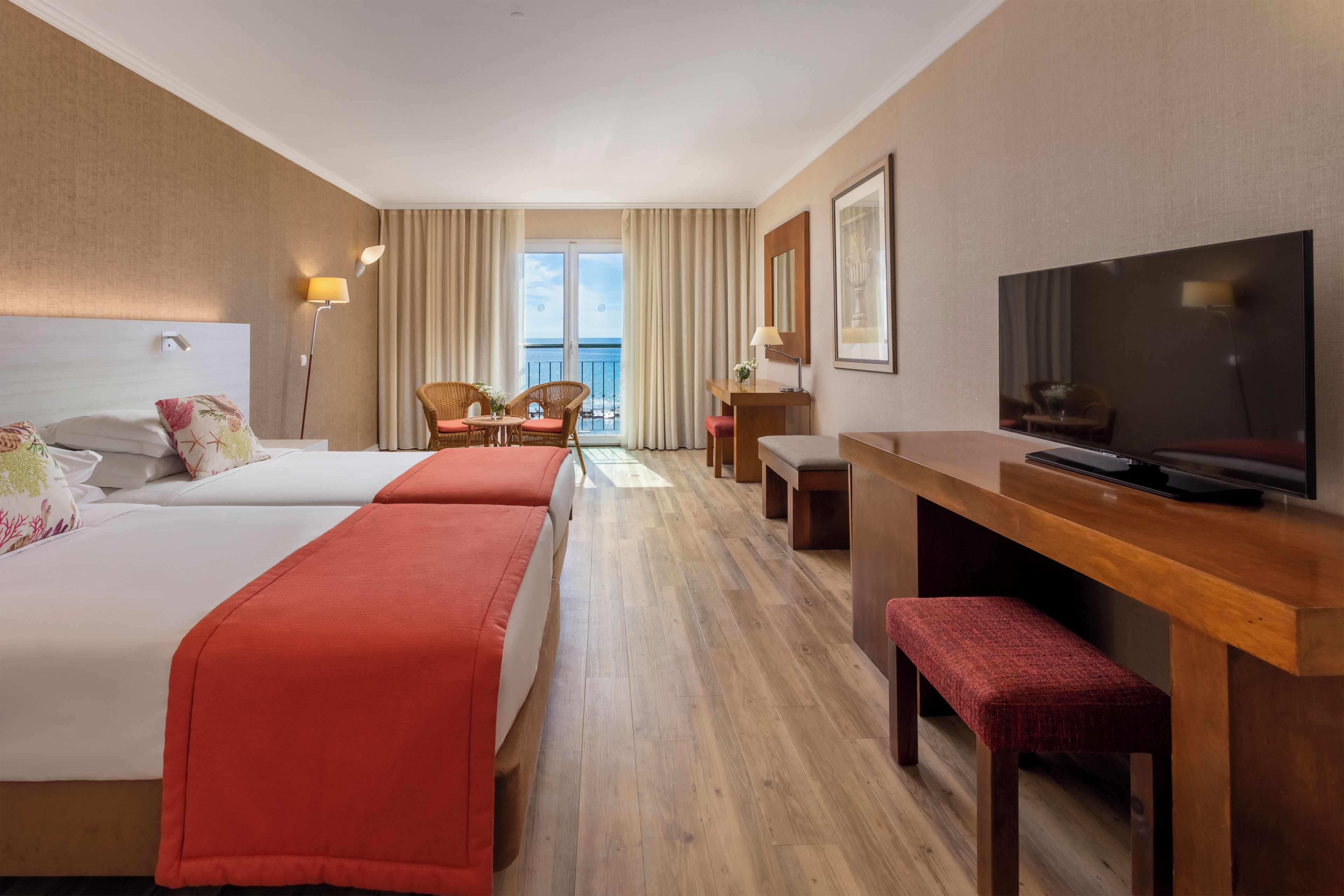 A hotel room with two beds, a desk, a television, and a balcony with a view of the sea.
