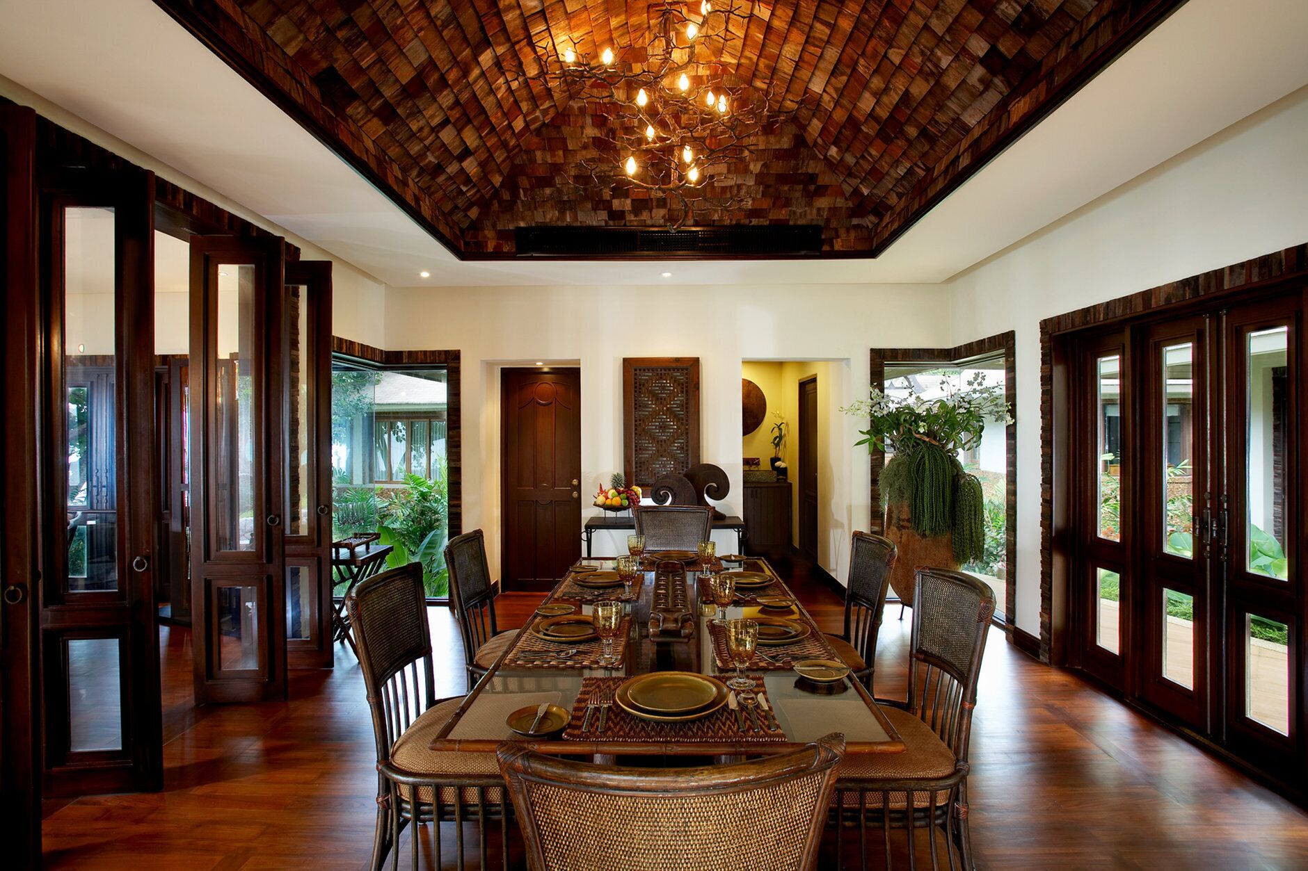 A spacious dining area with a long table set for a meal, surrounded by chairs. The room features a high wooden ceiling with a unique design, large windows on one side, and a view of greenery outside.