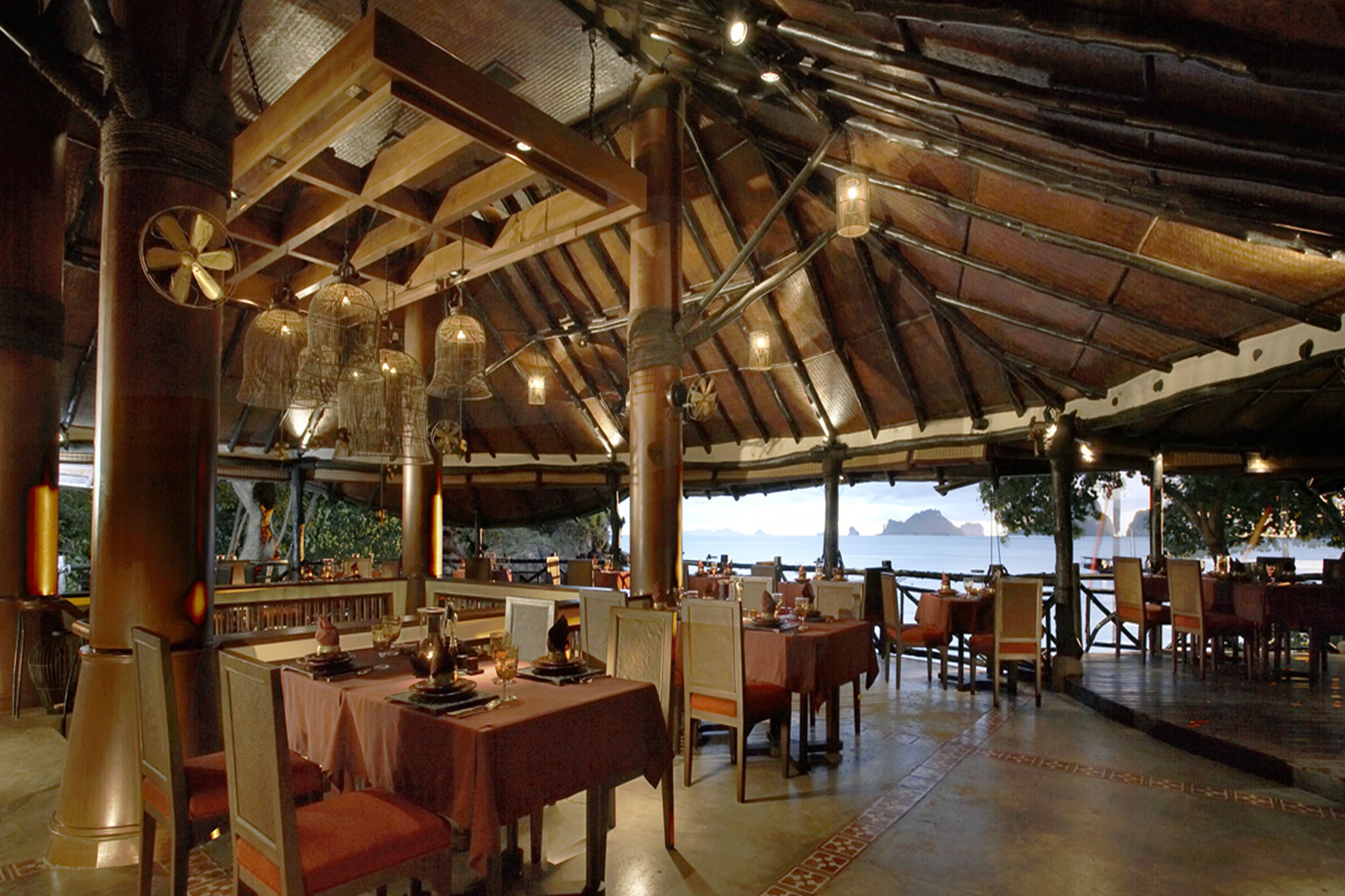 Een eethoek binnen met houten balken en hangende lampen. Tafels staan klaar met stoelen, en er is uitzicht op de zee door grote ramen.