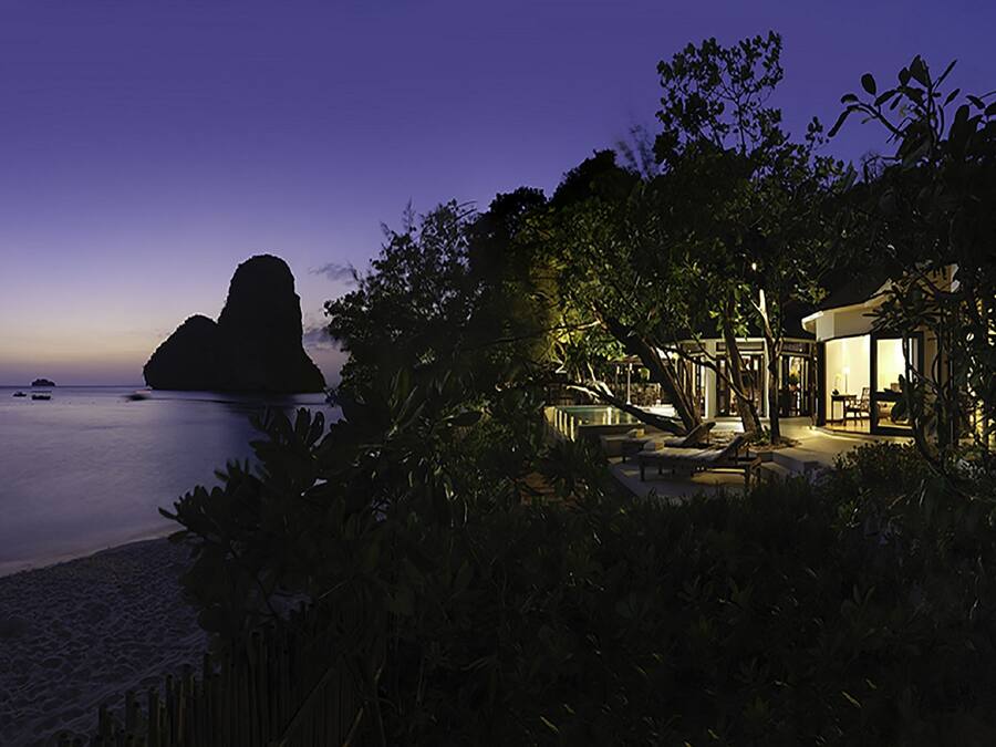 Una proprietà fronte spiaggia al crepuscolo con una grande formazione rocciosa all'orizzonte, una piscina e una zona relax ben illuminata.