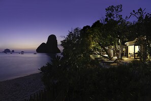 A beachfront property at dusk with a large rock formation in the distance, a pool, and a well-lit lounge area.