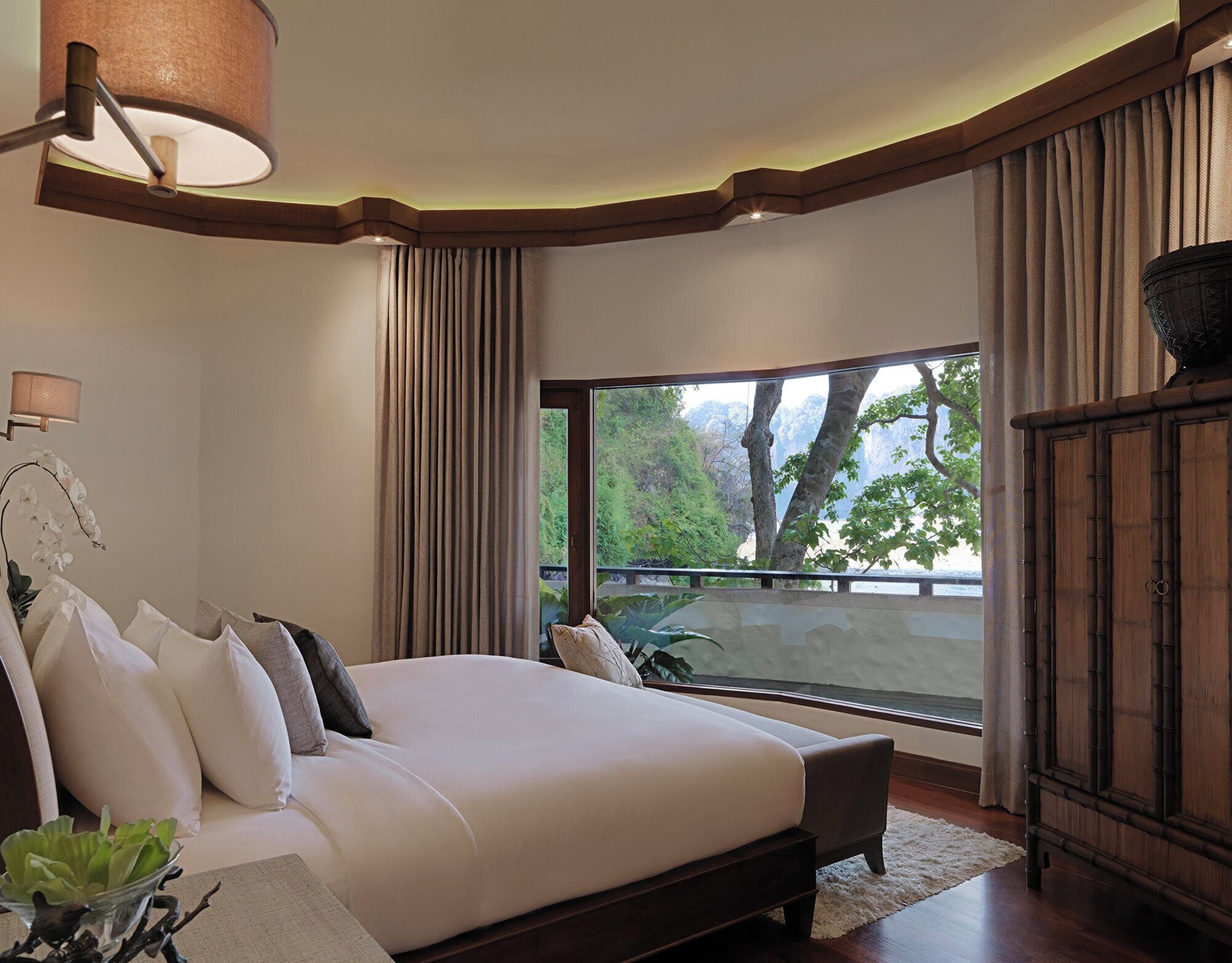 A bedroom with a large bed, a nightstand, a lamp, a view of a natural landscape through a window, and a wooden cabinet.