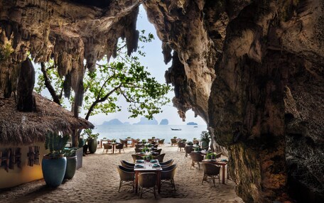 An outdoor dining area with tables and chairs, a thatched-roof building, and a view of the sea.