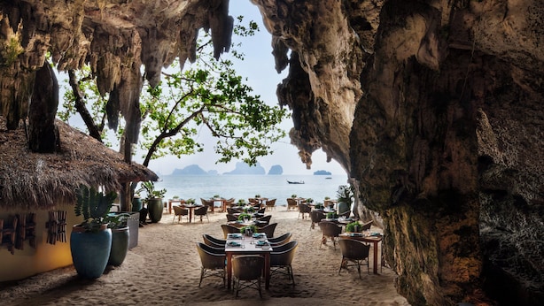 An outdoor dining area with tables and chairs, a thatched-roof building, and a view of the sea. - Rayavadee (Krabi)