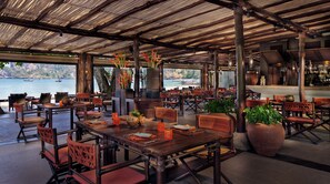 An outdoor dining area with wooden tables and chairs, large potted plants, and a view of the water.