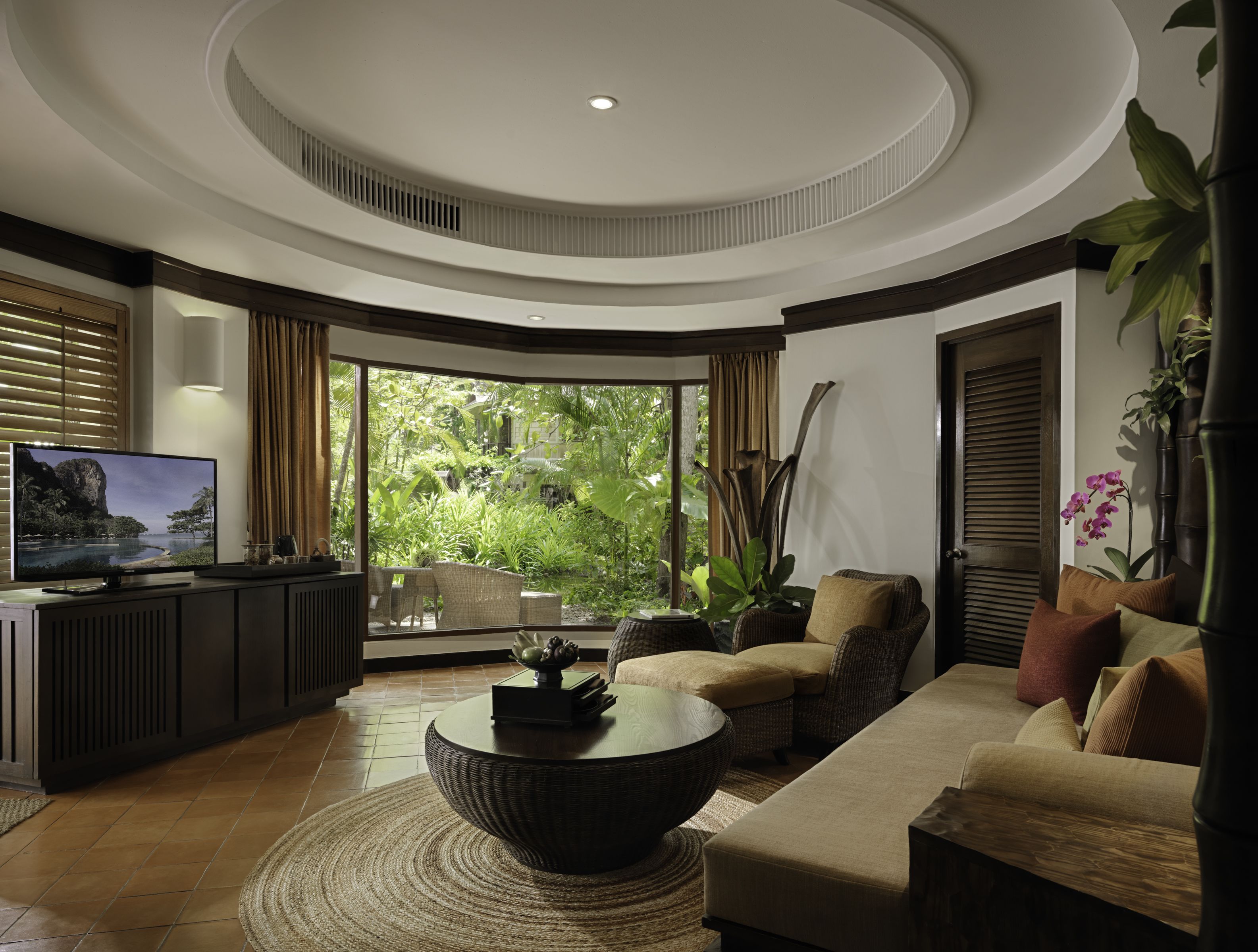 Een moderne woonkamer met een grote televisie, comfortabele zithoek, een ronde salontafel en uitzicht op weelderige begroeiing door grote ramen.