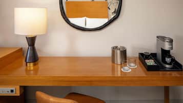A hotel room with a wooden desk, a lamp, a coffee maker, a cup, and a mirror.