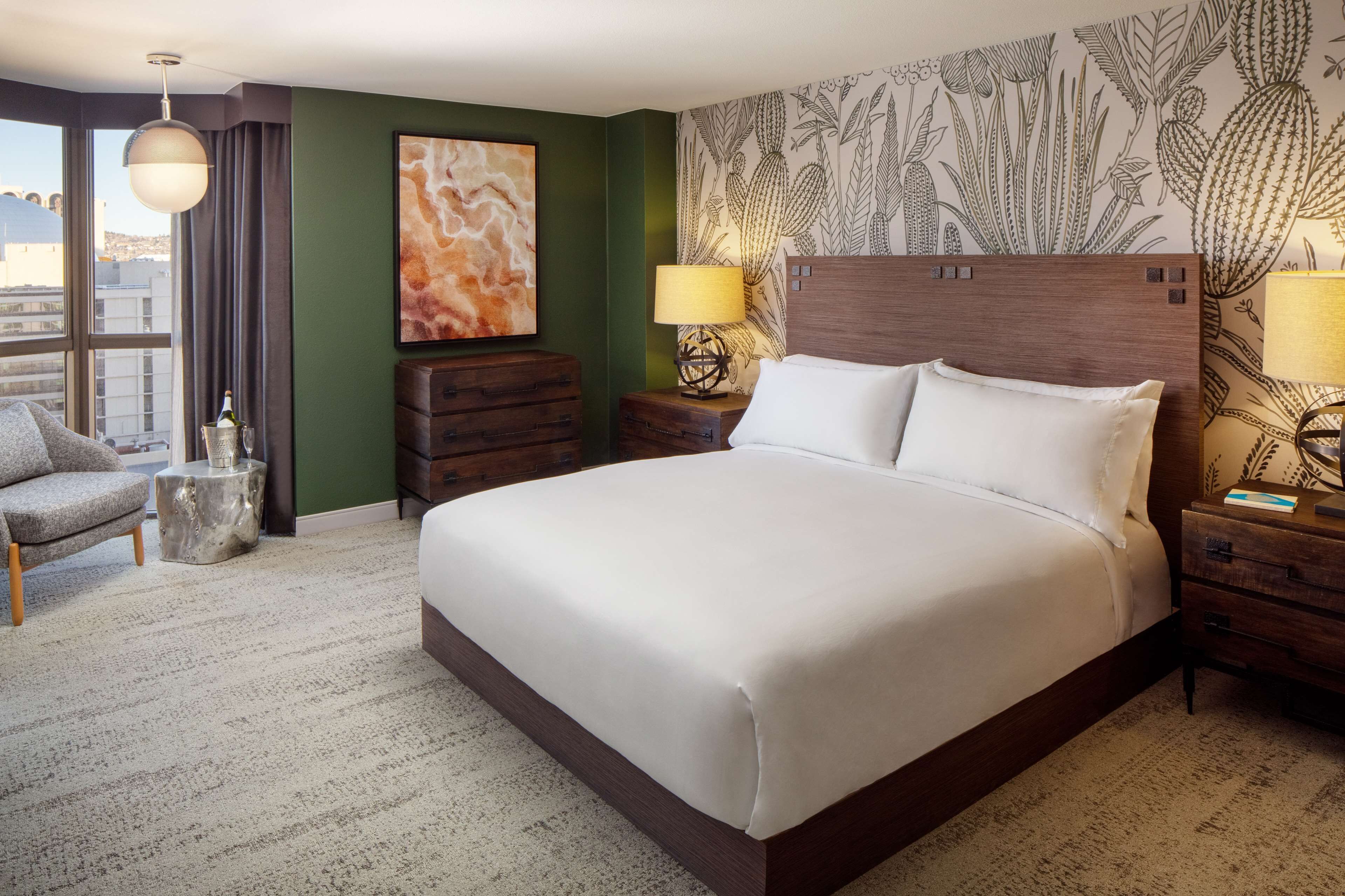 A hotel room with a large bed, two bedside tables with lamps, a chair, a small side table with a bottle, and a wall with a cactus pattern.