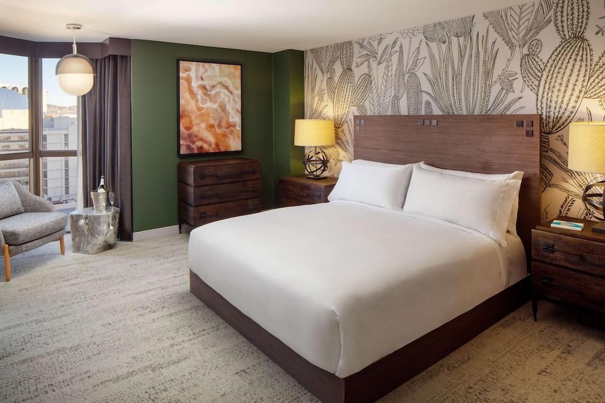 Habitación de hotel con una cama grande, dos mesitas de noche con lámparas, una silla, una pequeña mesita auxiliar, una pared decorativa con un motivo de cactus y un ventanal con cortinas.