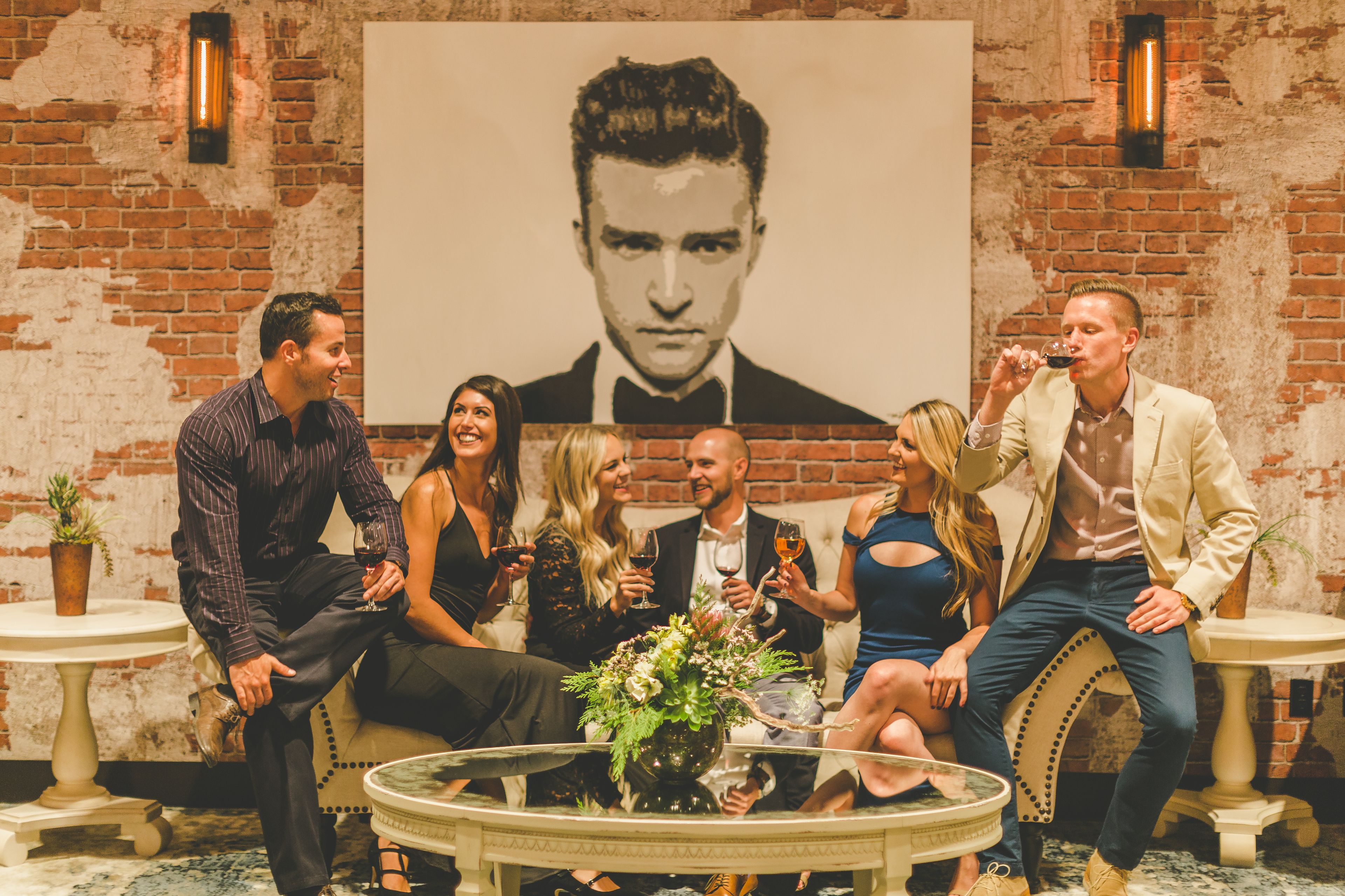 A group of people seated on a round table with a vase of flowers, in a room with brick walls and two small tables with potted plants.