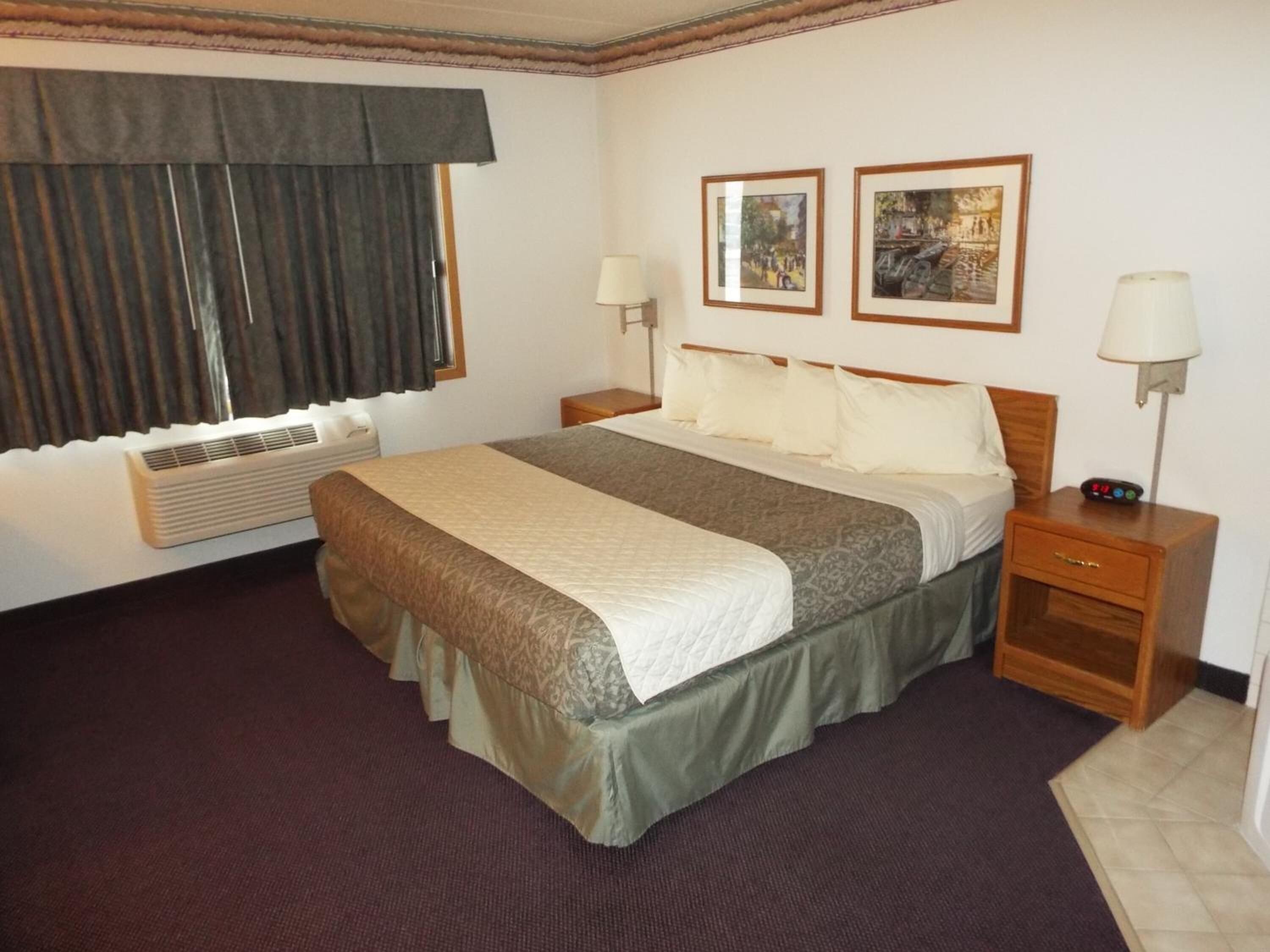 Quarto de hotel com uma cama grande, duas mesas de cabeceira, uma janela com cortinas, ar-condicionado fixado na parede e duas quadros emoldurados sobre a cama.