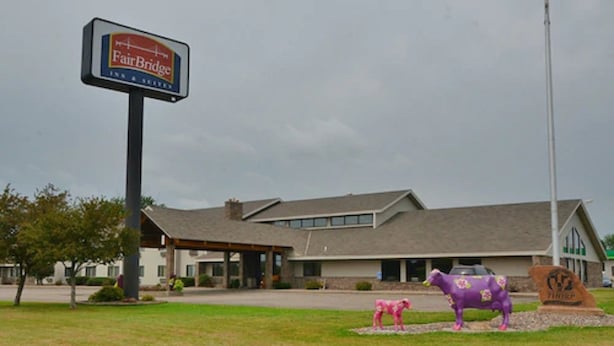 Un bâtiment de l’hôtel Fairbridge Inn & Suites, avec une grande enseigne, une entrée couverte et deux statues de chevaux décoratives en premier plan.