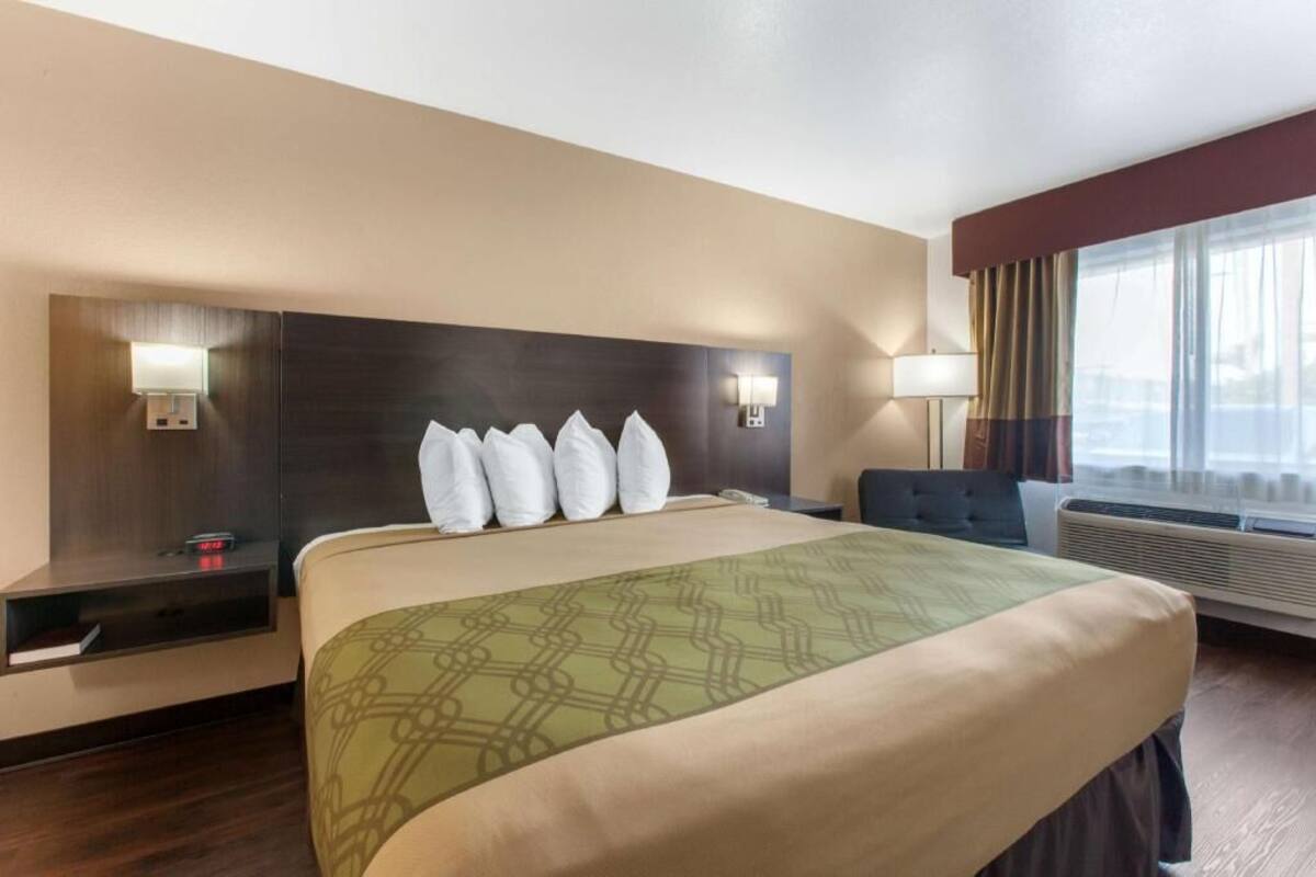 Ein Hotelzimmer mit einem großen Bett, Nachttischen, einem Schreibtisch, einem Sessel, einem Fenster mit Vorhängen und einer Wand-Klimaanlage.
