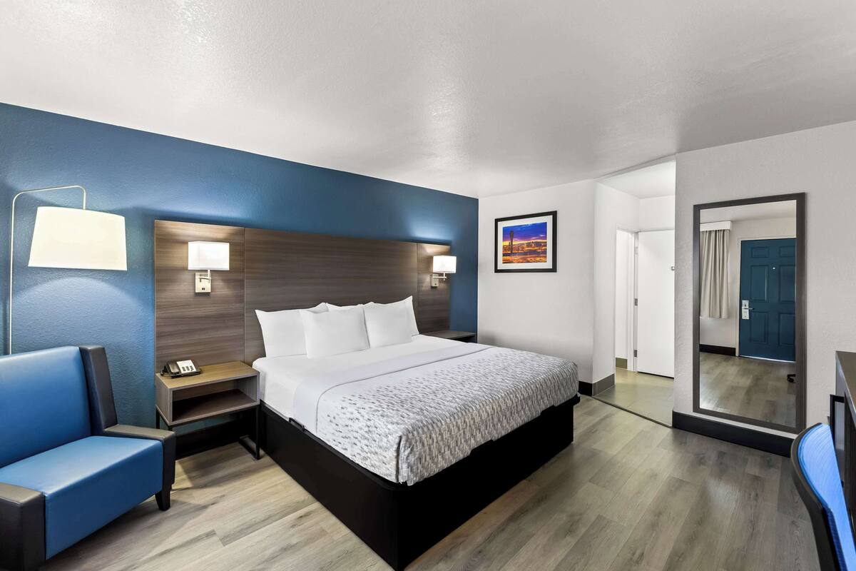 Ein modernes Hotelzimmer mit einem großen Bett, einem Nachttisch mit Lampe, einer kleinen Sitzecke und einem großen Spiegel.