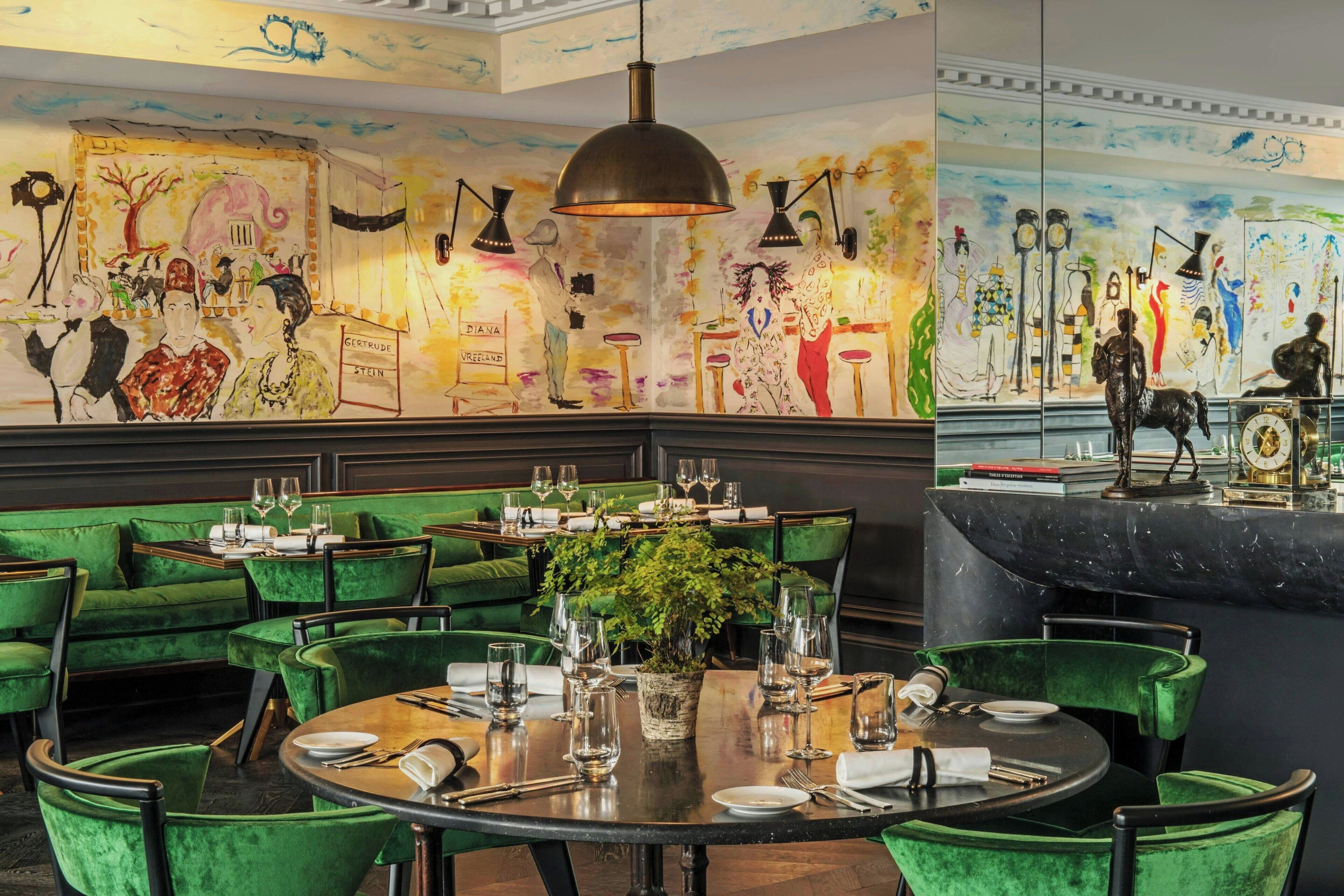 A restaurant with green velvet seating, a round table with wine glasses, and a mural with various figures and text.