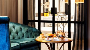 A room with a green upholstered sofa, a round table with a teapot and fruit, and a large window with curtains.