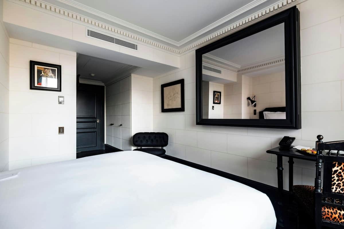 A hotel room with a large bed, a black-framed mirror, a small table with a phone and fruit, and a leopard-print chair.