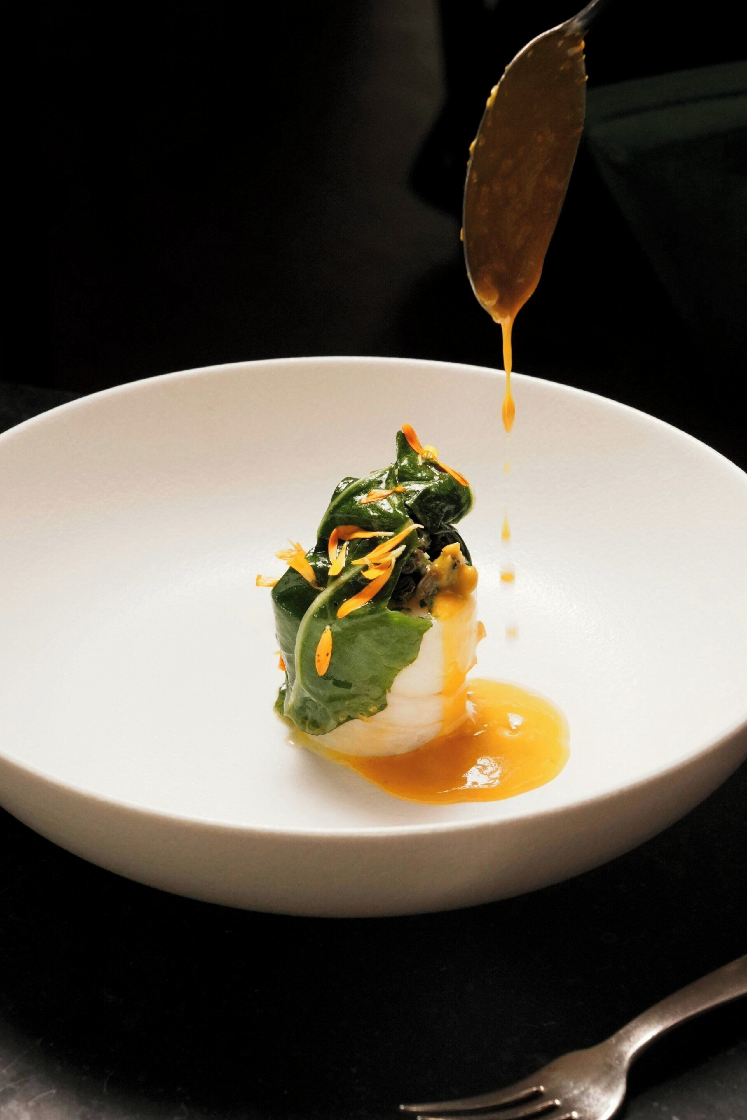 A white plate with a dish of greens and a sauce, a fork, and a spoon on a dark surface.