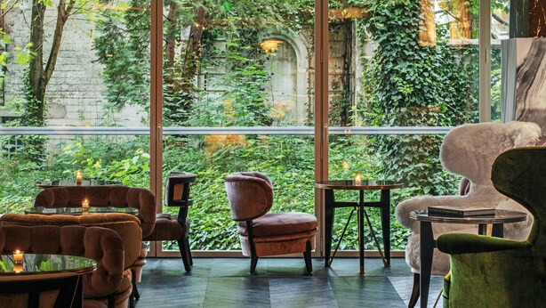 A cozy indoor seating area with plush chairs and tables, large windows offering a view of lush greenery and an old stone wall.