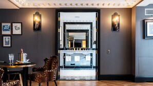 A hotel lobby with a wooden herringbone floor, leopard print chairs, a round table with a pitcher and glasses, and wall-mounted sconces.