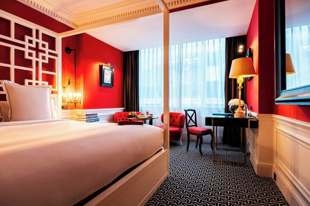 A hotel room with a bed, a desk, a chair, a lamp, and a view of the outside through large windows.
