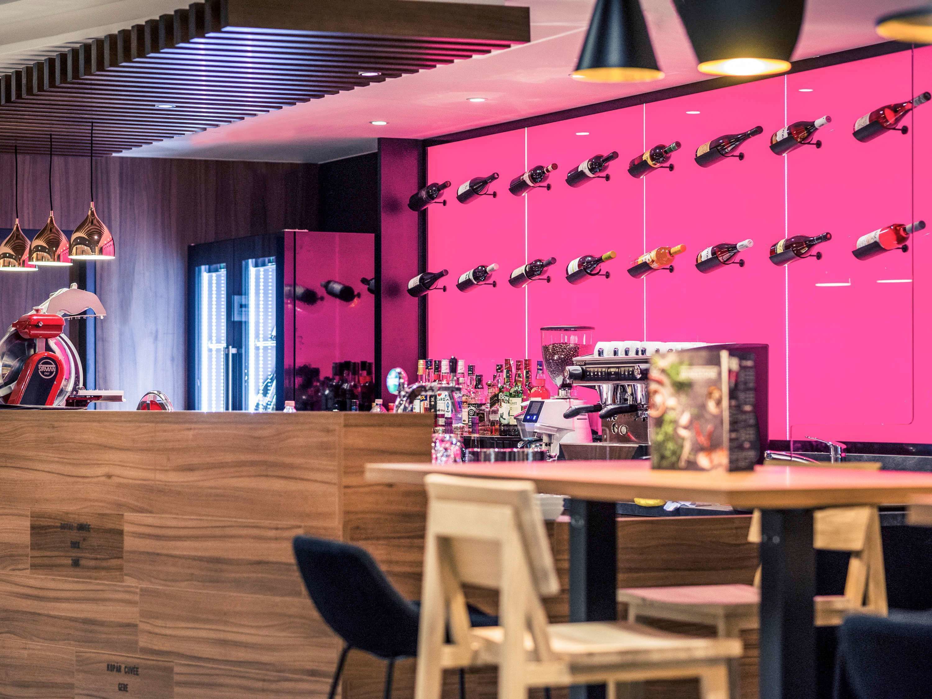 A bar area with a modern design, featuring a pink wall with hanging bottles, a coffee machine, and a variety of liquor bottles.