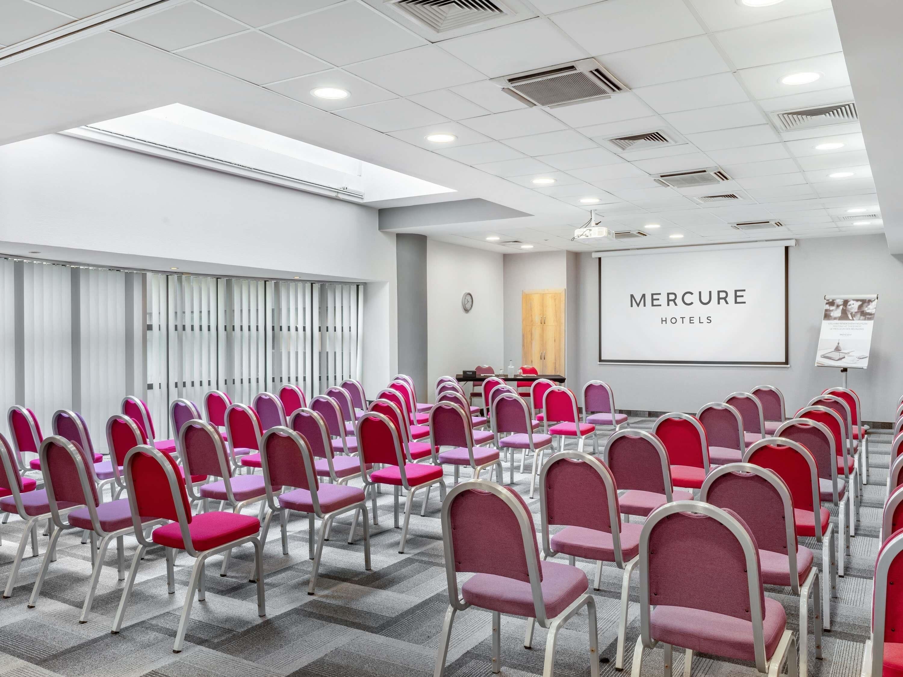Una sala conferenze con file di sedie rosse, uno schermo di proiezione e un cartello con la scritta 'Mercure Hotels'.