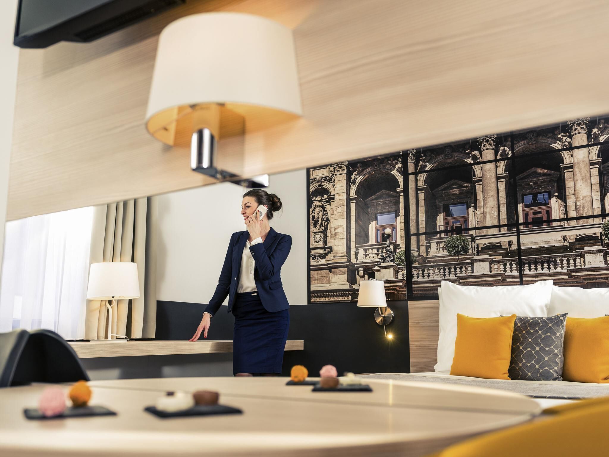 Habitación de hotel con un gran ventanal, cama con almohadas amarillas y grises, escritorio con lámpara y una pared decorativa que muestra una vista de un edificio clásico.
