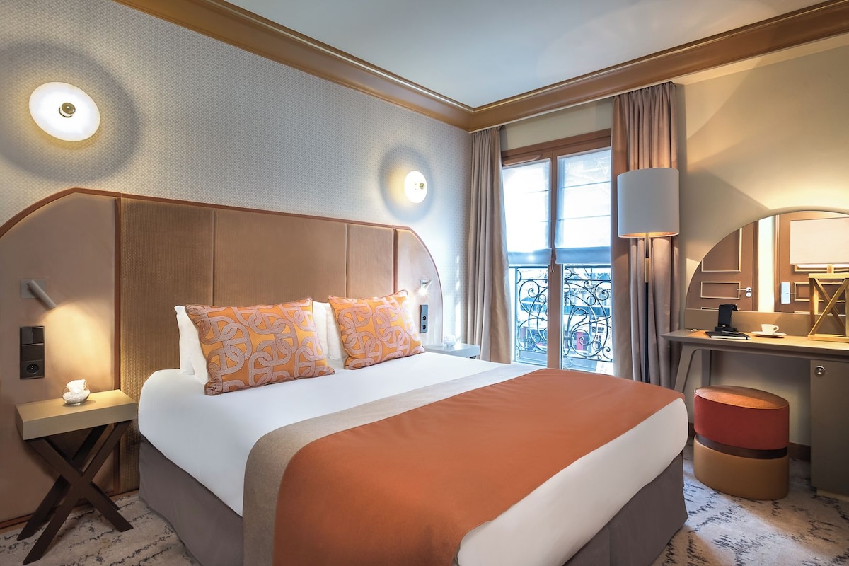 A hotel room with a large bed, a desk, a chair, a lamp, and a window with curtains.