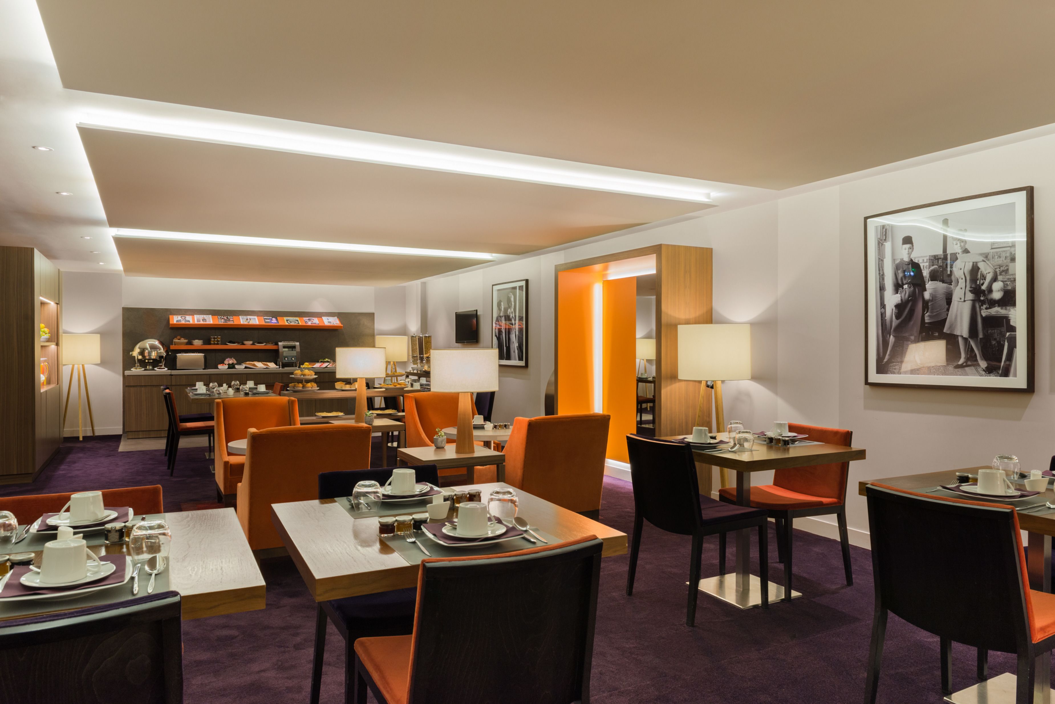 Ein Essbereich eines Hotels mit gedeckten Tischen, eine Küche im Hintergrund und ein Gang zu einem weiteren Raum.