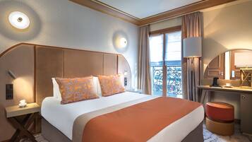 A hotel room with a large bed, a desk, and a chair. There is a window with curtains, a lamp, and a nightstand with a phone.