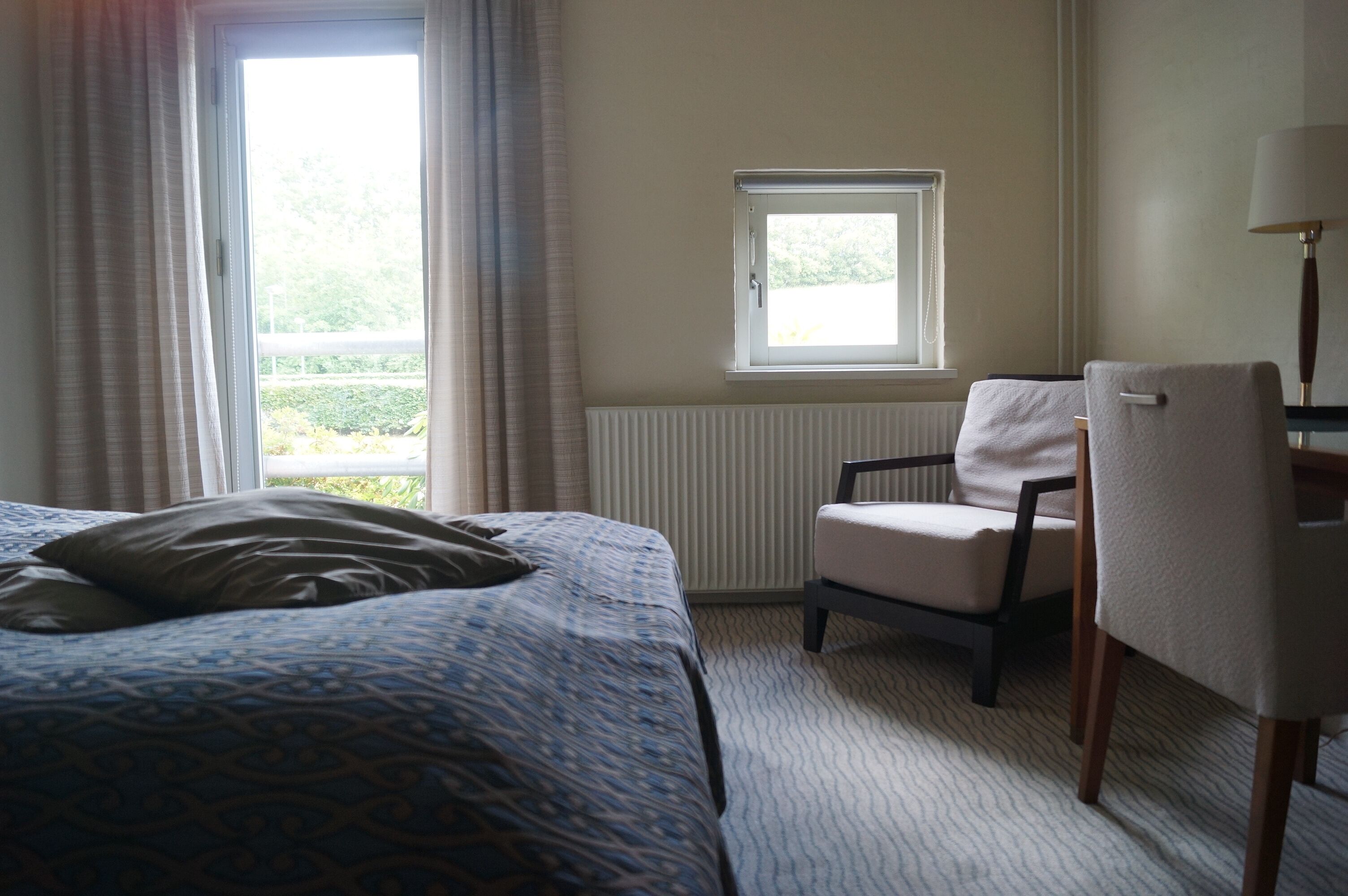 A hotel room with a bed, a chair, a window, and a radiator.