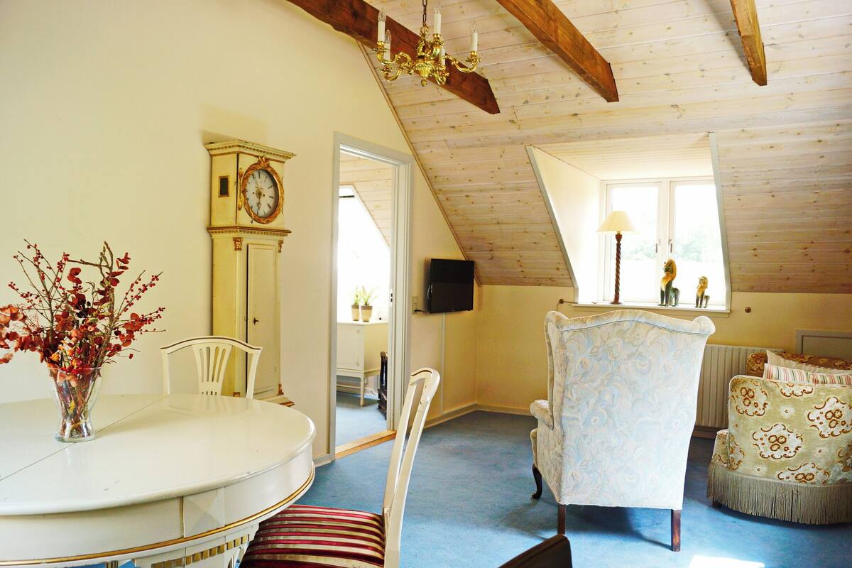 A room with a dining table, chairs, a clock, a television, and a window with a view.