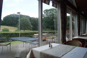 Un área de comedor al aire libre con mesas y sillas, amplias ventanas que ofrecen vistas a un jardín ajardinado y árboles.