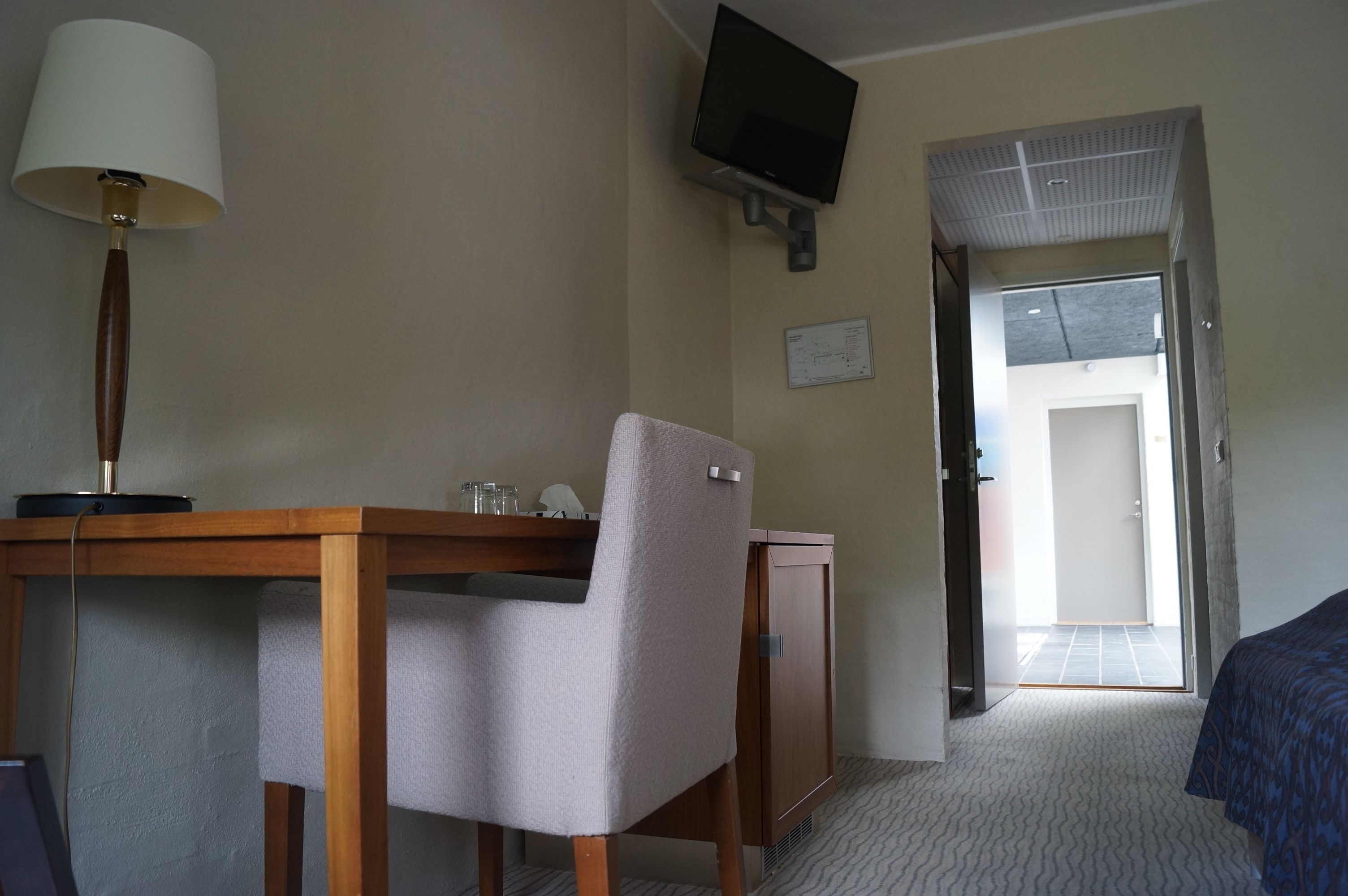 Habitación de hotel con un escritorio de madera, una silla, un televisor montado en la pared y una puerta que da a un balcón.
