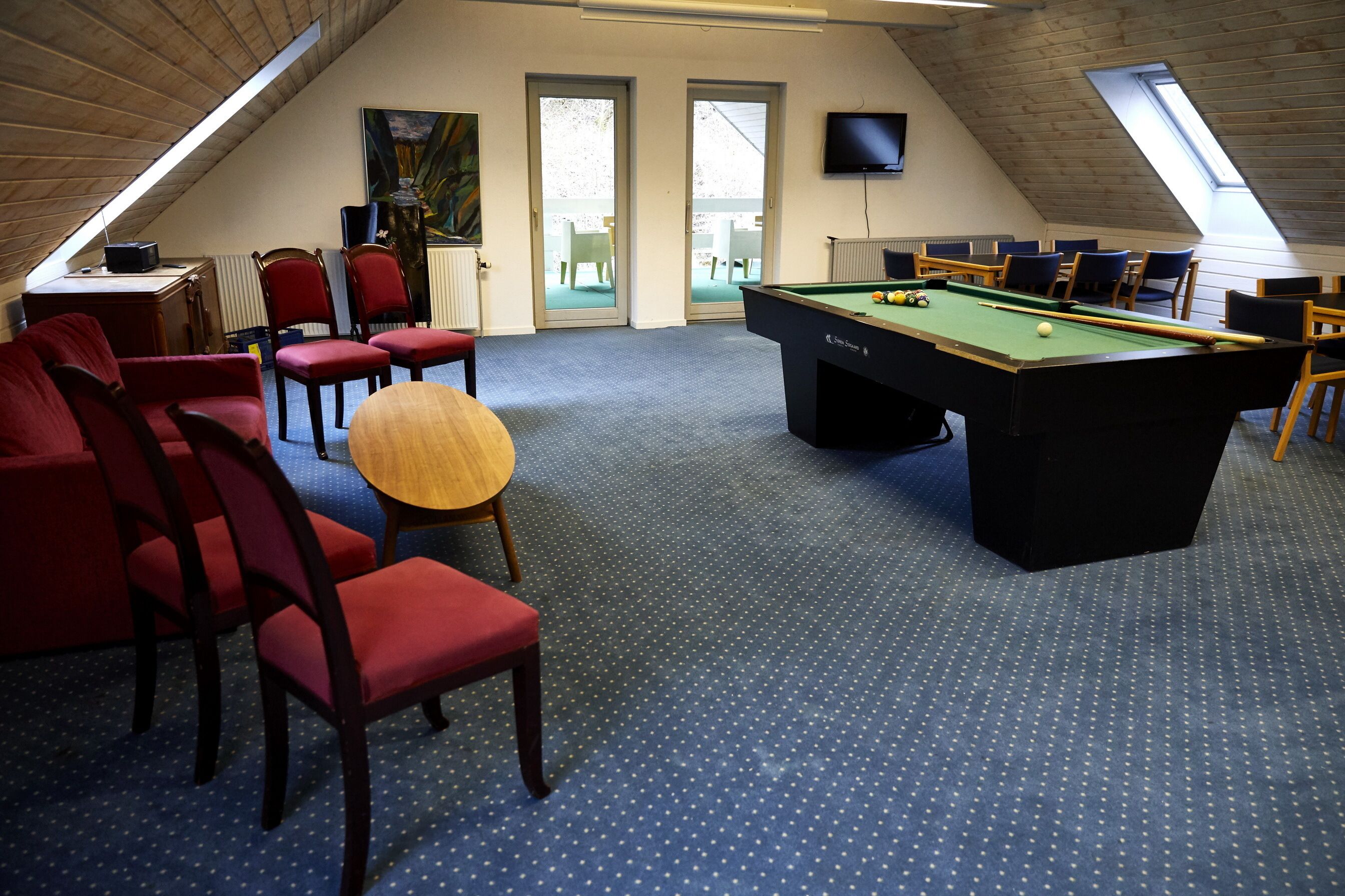 A room with a pool table, red chairs, a round wooden table, and a television on the wall.
