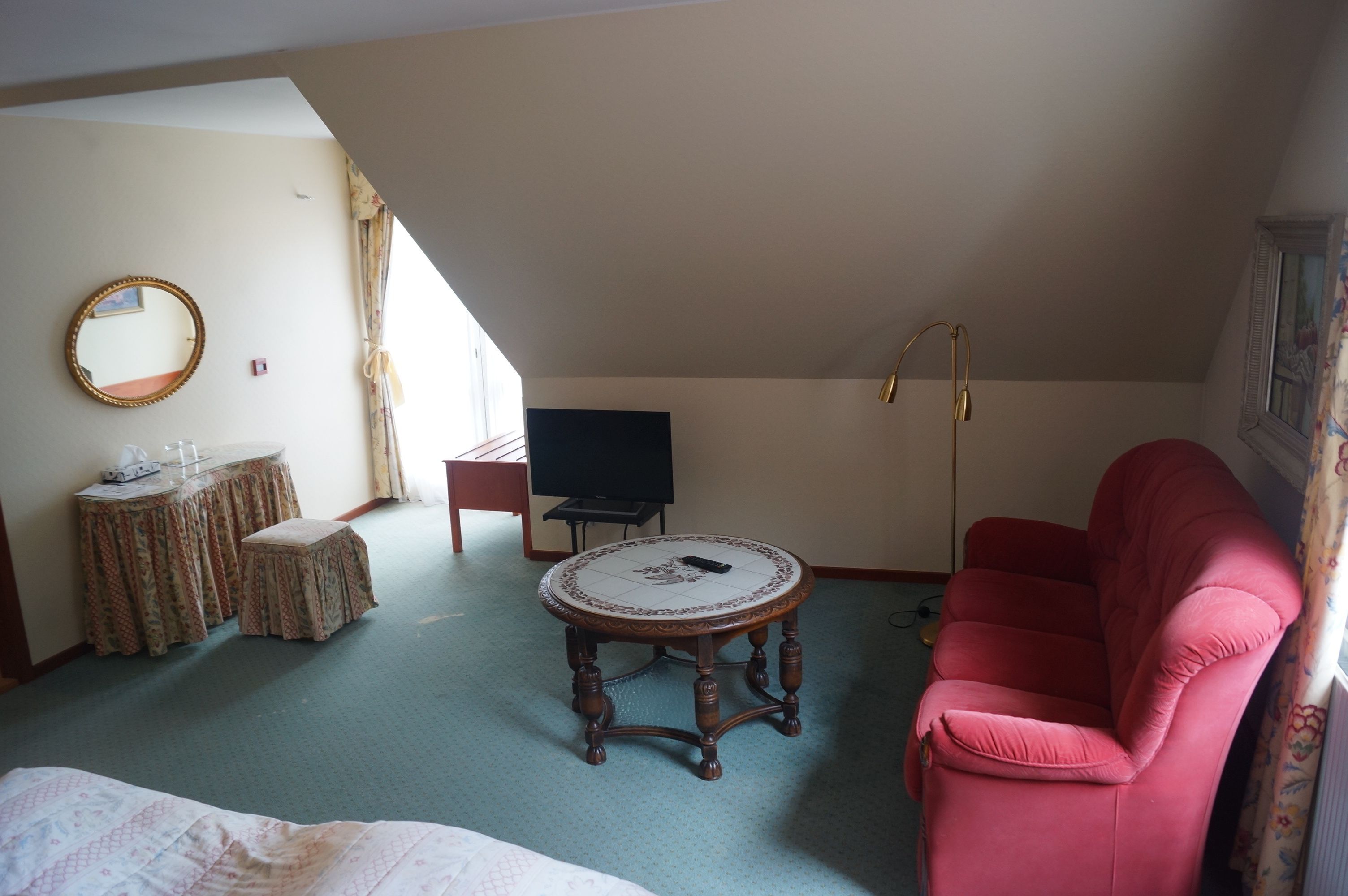 Habitación de hotel con una cama, un sofá rojo, una mesita redonda pequeña, un televisor, un espejo y una ventana con cortinas.