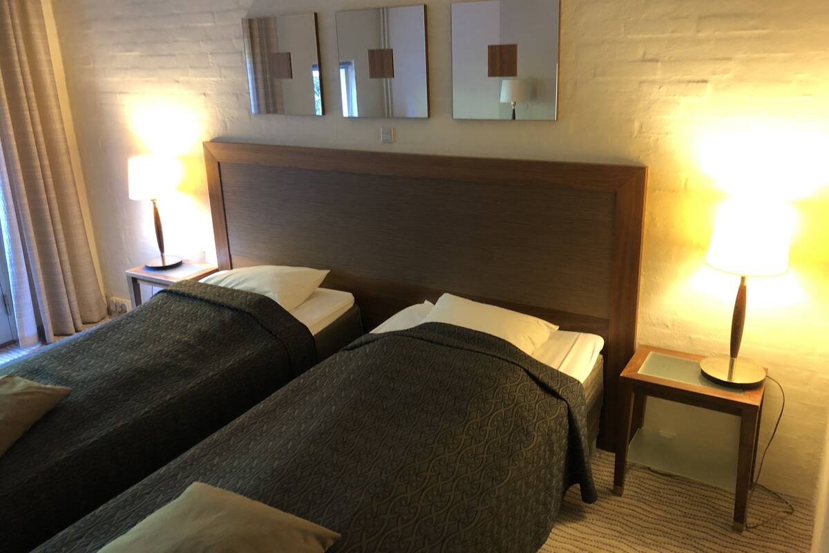 A hotel room with two single beds, a wooden headboard, a nightstand with a lamp, and a wall-mounted mirror.
