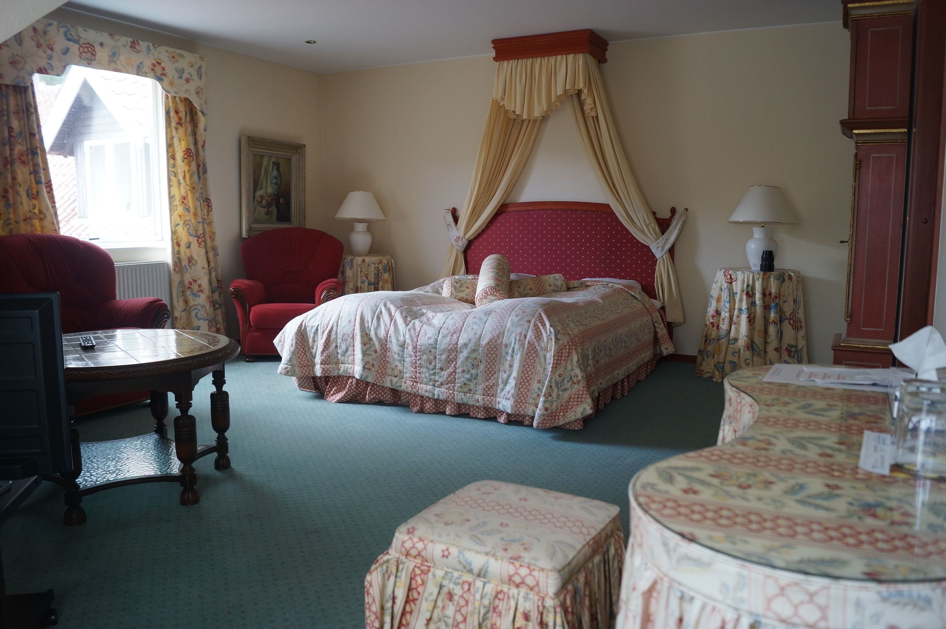 A bedroom with a four-poster bed, a red armchair, a round table, and a small table with a lamp.
