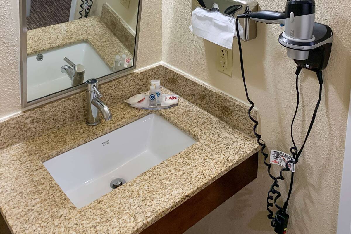 A bathroom with a granite countertop sink, a mirror, a towel dispenser, and a hair dryer mounted on the wall.