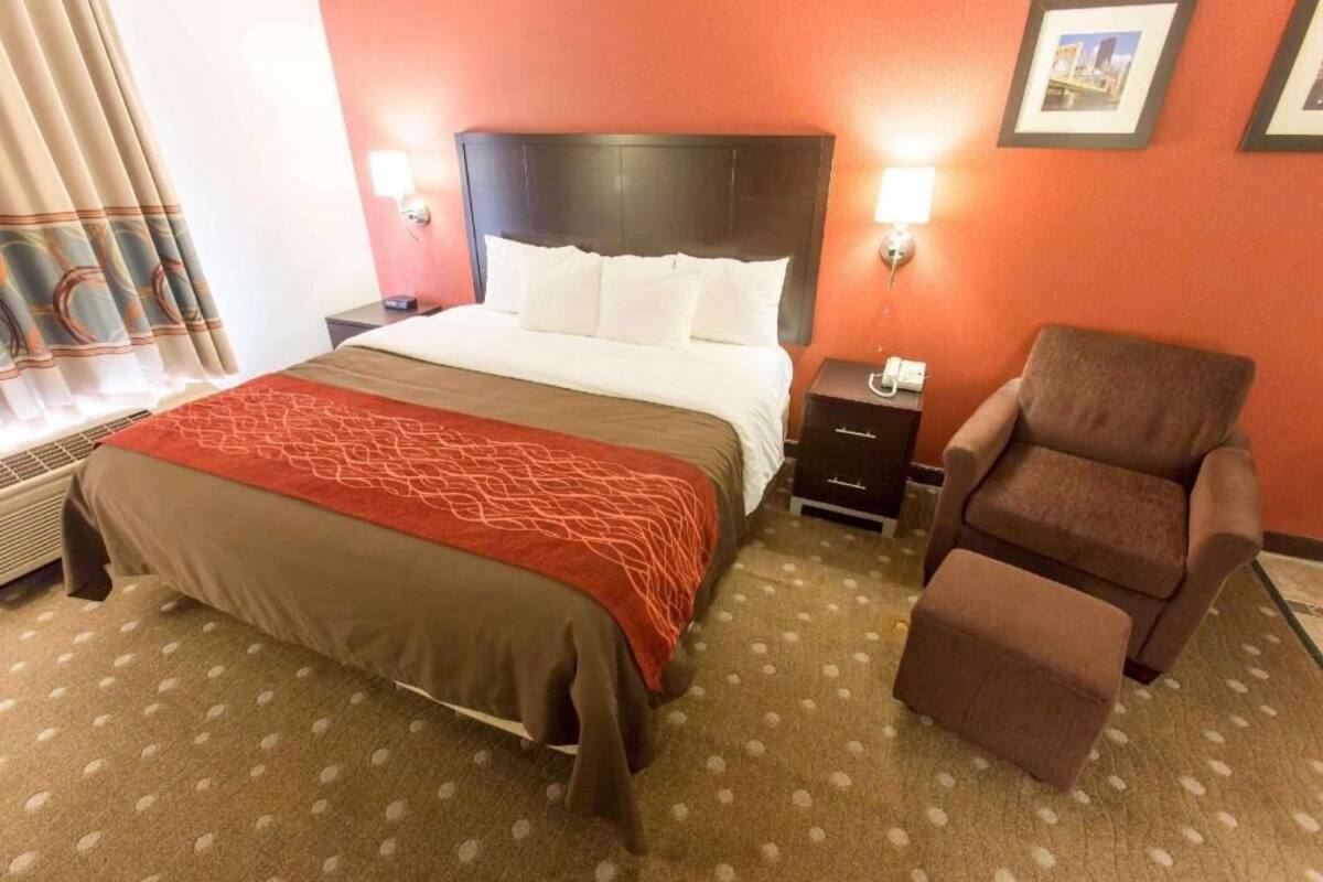 A hotel room with a bed, a chair, a nightstand, and a wall-mounted lamp.