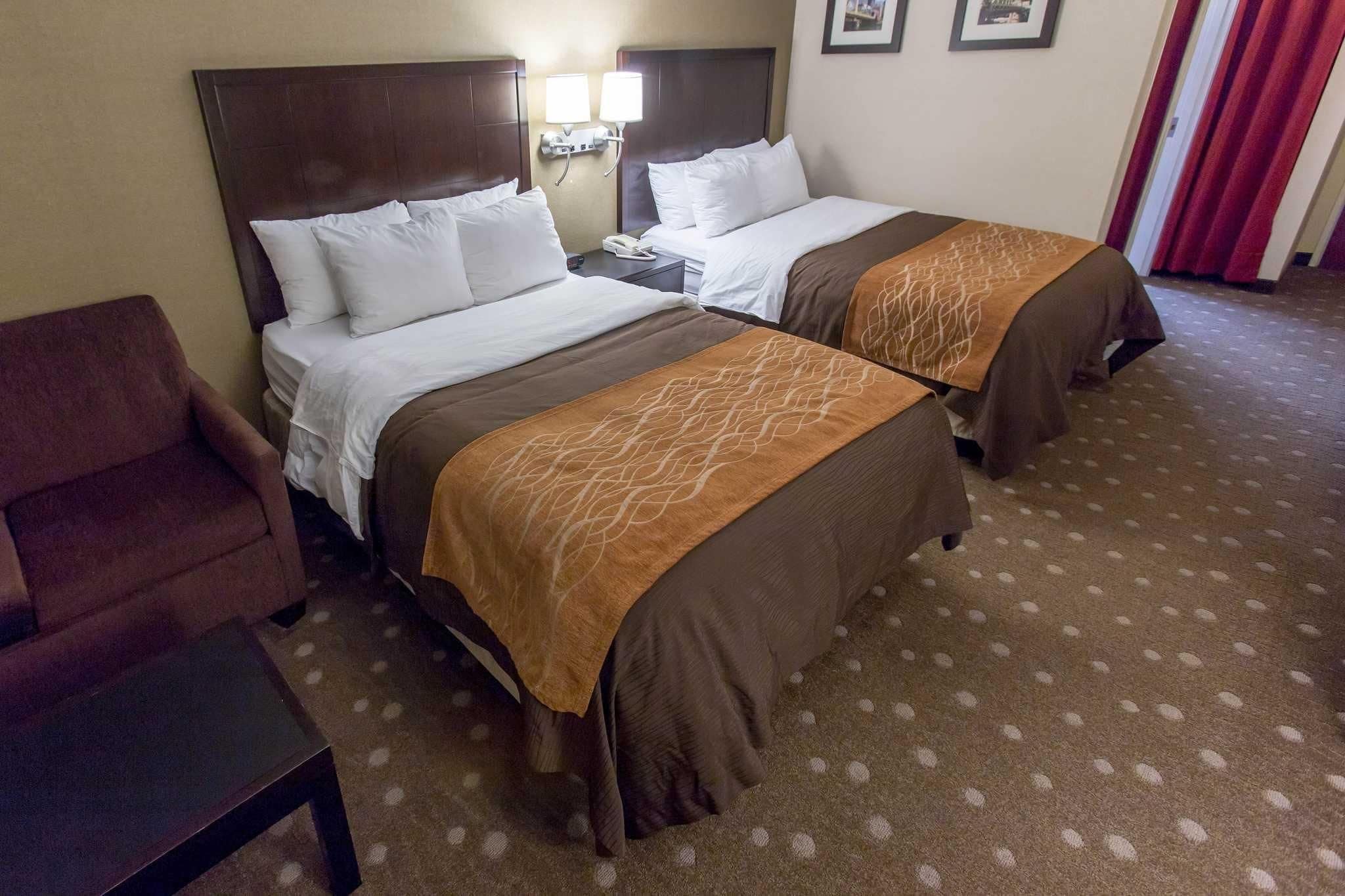 A hotel room with two beds, a brown armchair, a small table, and a carpeted floor.