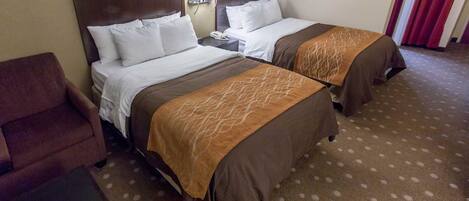 A hotel room with two beds, a brown armchair, a small table, and a carpeted floor.
