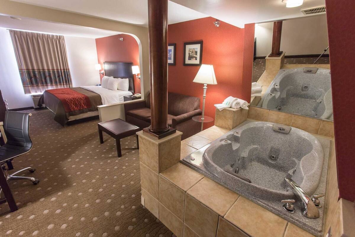 A hotel room with a bed, a desk, a chair, a sofa, a lamp, and a large bathtub.